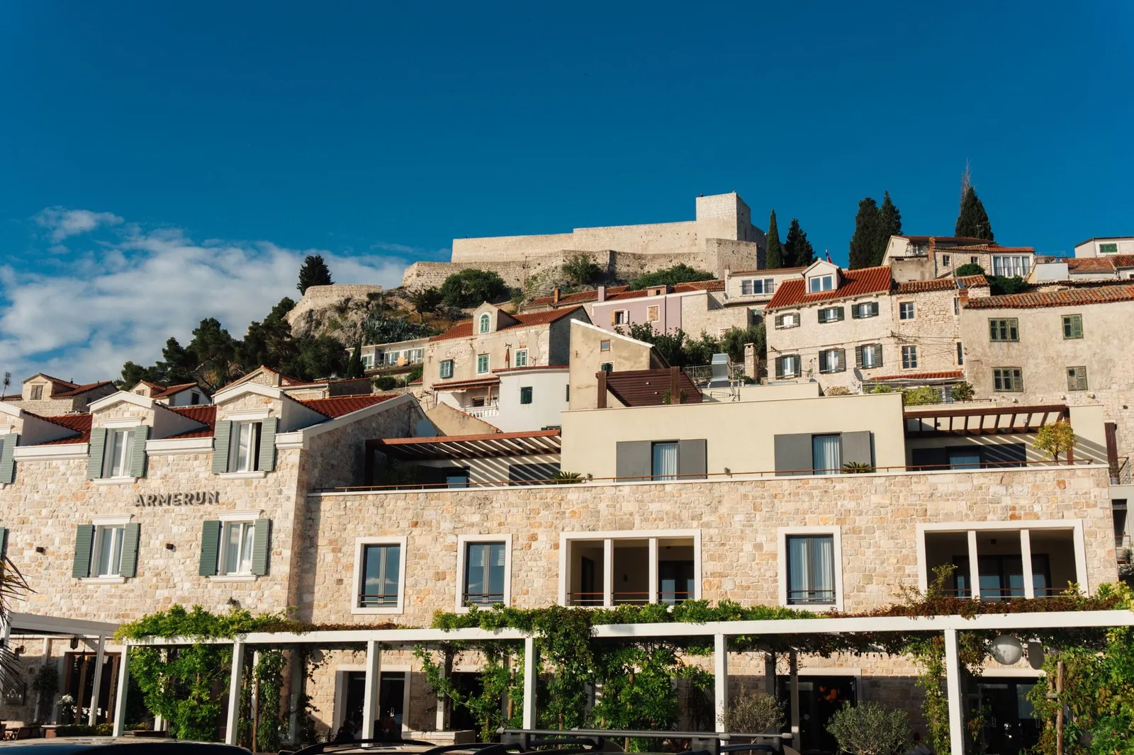 Scenic view of historic stone buildings in Hvar, Croatia with a fortress in the background under a clear blue sky.