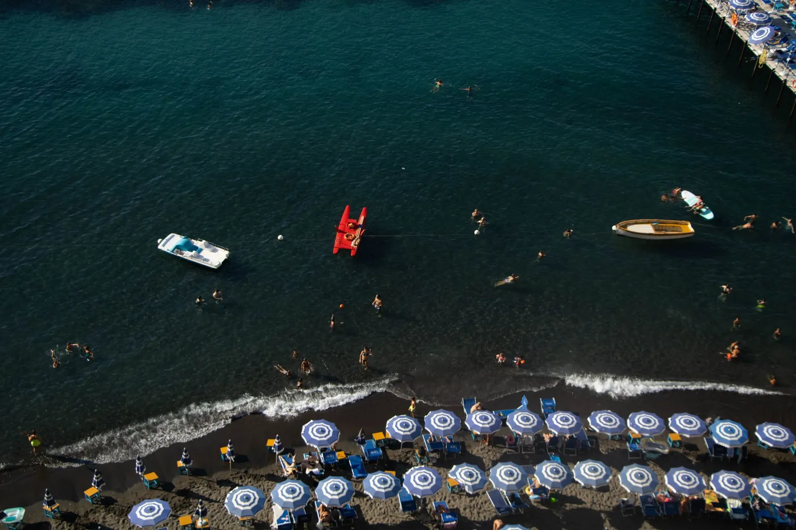A picturesque aerial view of Sorrento beach featuring blue umbrellas, swimmers, and boats under a summer sun.