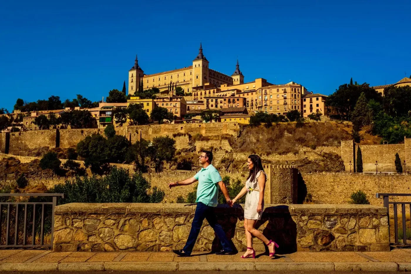 A couple holding hands walks near the historic Alcazar of Toledo, Spain on a sunny day.